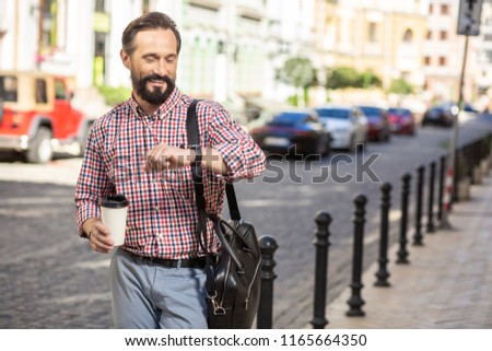 In pace with time. Cheerful handsome man looking at his wristwatch while walking along the street