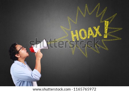 Young Asian man wearing white and blue shirt shouting using megaphone, angry expression. Close up body portrait. With a hoax sign Royalty-Free Stock Photo #1165765972