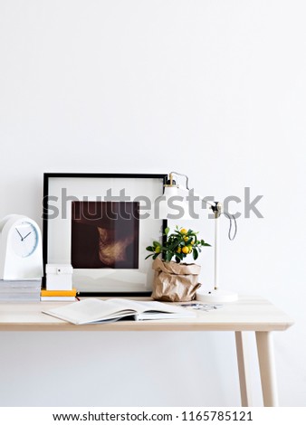 Interior shot of working table with tiny citrus tree and lamp on top in composition with book and painting against white wall.