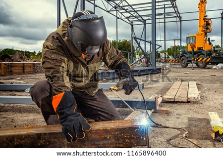 A young  man welder in brown uniform, welding mask and welders leathers, weld  metal  with a arc welding machine at the construction site, blue sparks fly to the sides Royalty-Free Stock Photo #1165896400