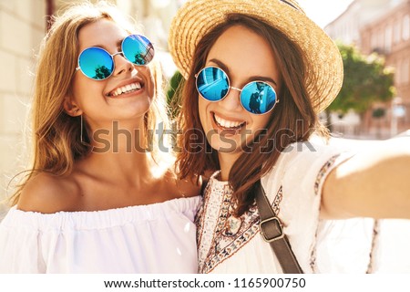 Two young female smiling hippie brunette and blond women models in summer white hipster dress taking selfie photos for social media on smartphone on the street background. Surprise face, emotions,