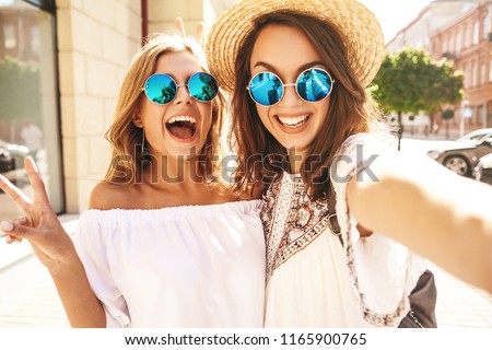 Two young female smiling hippie brunette and blond women models in summer white hipster dress taking selfie photos for social media on smartphone on the street background. Surprise face, emotions, Royalty-Free Stock Photo #1165900765