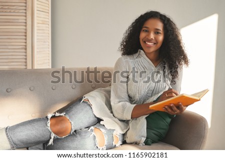 Horizontal portrait of charming positive young mixed race female in stylish shirt and ripped blue jeans smiling broadly, lying on couch with diary, planning vacations, feeling happy and inspired