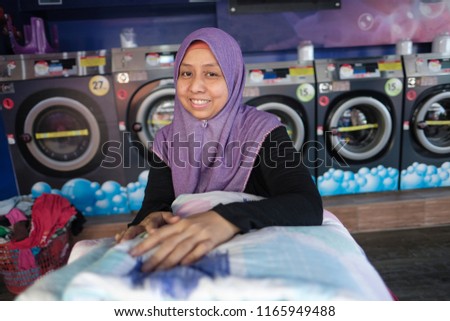 A young malay mother wearing hijab smiling happily sending her clothes to a self-service laundry shop