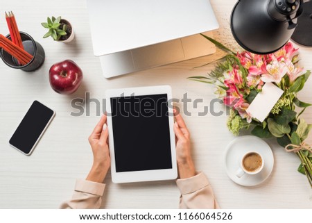 partial view of female freelancer holding digital tablet with blank screen at table with coffee cup, gadgets and flowers with greeting card 