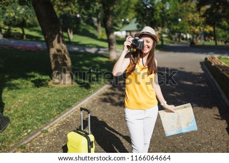 Joyful traveler tourist woman in hat with suitcase city map take pictures on retro vintage photo camera in city outdoor. Girl traveling abroad to travel on weekend getaway. Tourism journey lifestyle