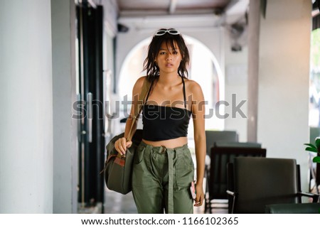 An attractive and young Chinese Asian millennial teenager girl walks down an corridor in the a city during the day. She is wearing a sling bag on her shoulder and is wearing street wear clothes. 