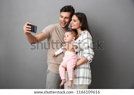 Portrait of a happy young family with their little baby girl isolated over gray background, taking a selfie