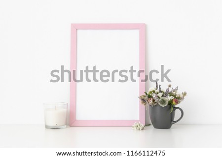 Pink portrait frame mockup with dried field wild flowers in gray mug and white candle on white wall background. Empty frame, poster mock up for presentation design. Template frame for text, lettering