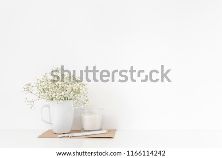 Background with stationary, white candle and bouquet of white flowers in mug on white wall background, elegant soft home decor. Copy space for text. Empty space for lettering, text.