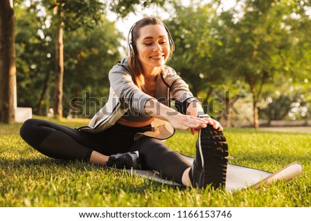 Image of smiling fitness woman 20s wearing headphones working out and stretching legs while sitting on exercise mat in green park