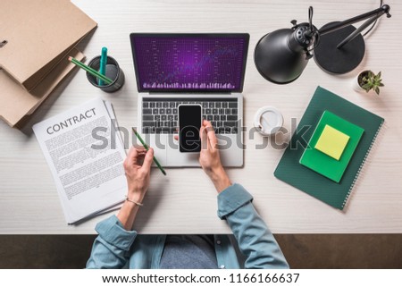 cropped image of businessman with smartphone at table with contract, stationery and laptop with graph on screen
