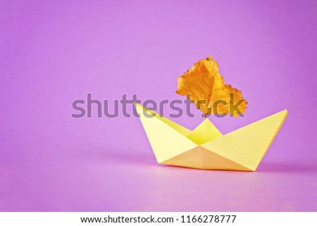 Yellow paper boat from origami with an autumn leaf on a purple background.