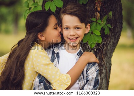 Two little kids in love. Portrait of beautiful little girl hugging sweet boy and kissing him on cheek. Tree in background.