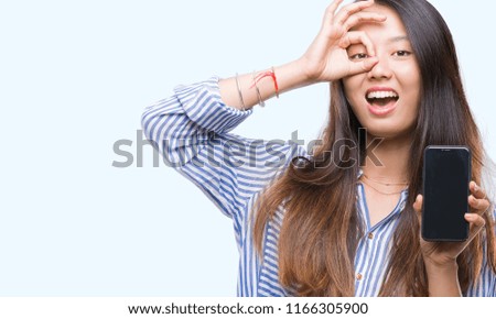 Young asian woman showing blank screen of smartphone over isolated background with happy face smiling doing ok sign with hand on eye looking through fingers