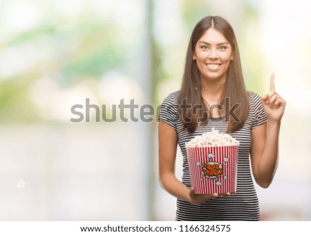 Young beautiful hispanic eating popcorn surprised with an idea or question pointing finger with happy face, number one