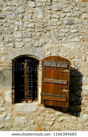 A rustic shuttered window with ornate security bars set in a stone wall in Pujols, Lot-et-Garonne, France. This historic village is a member of "Les Plus Beaux Villages de France" association.