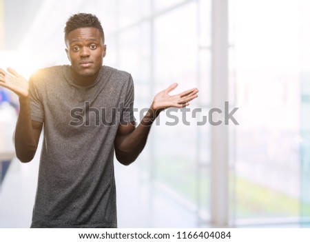 Young african american man wearing grey t-shirt clueless and confused expression with arms and hands raised. Doubt concept.