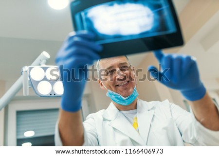 Smiling happy dentist holding x-rays. X-ray of a human jaw in the hands of the dentist. Close up male dentist pointing at patient's X-ray image in dental office.