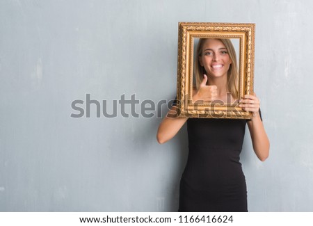 Beautiful young woman over grunge grey wall holding vintage frame happy with big smile doing ok sign, thumb up with fingers, excellent sign