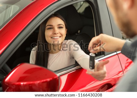 Beautiful and young woman sitting in red car and smiling. Female customer holding her hand and receiving keys of her new car from manager who working in modern car dealership.