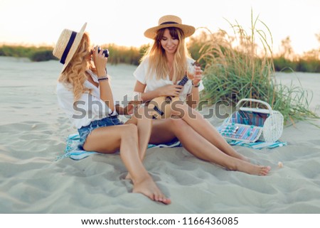 Two carefree happy girls  enjoying a beach party with music and picnic in summer evening. Blonde pretty girl playing ukulele guitar and her friend making picture .