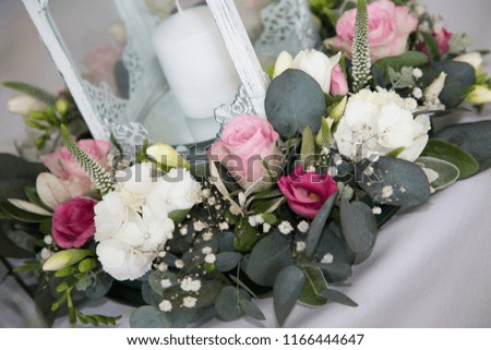 floral display on a wedding day table of pink roses and white carnations with green leaves around a vase with a white candle inside