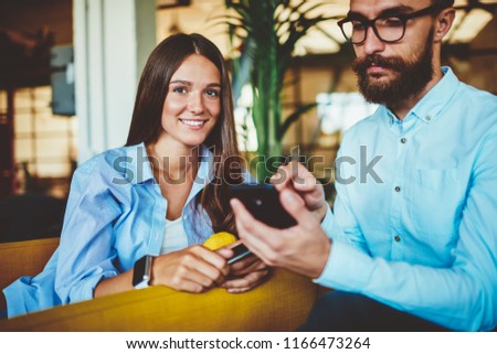 Portrait of successful female teenager smiling at camera while male friend sitting near and checking notification on telephone device using 4G internet, hipster guy sending sms message on smartphone