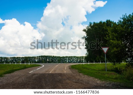 Road sign of give way on crossroads of dirt road before asphalt road. Landscape with highway and trees. White clouds in blue sky. Сareful driving. Traffic Laws.