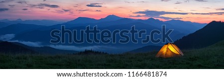 Glowing orange tent in the mountains under dramatic evening sky. Red sunset and mountains in the background. Summer landscape. Panorama