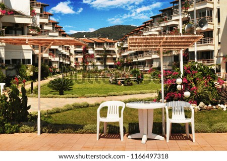 Hotel buildings and green park with grass, bushes, flowers, white plastic chairs and table with forest and blue sky with clouds on a background