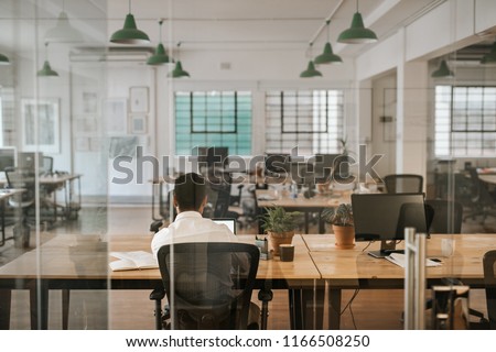 Rearview of a young businessman sitting alone at his desk in an office working on a laptop and going over paperwork Royalty-Free Stock Photo #1166508250