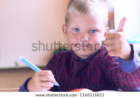 Happy smiling boy with thumb up sitting in schoolclass. Kid indoors of the class room with school desk on a background. Back to school. .