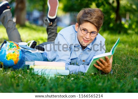 cute young boy in round glasses and blue shirt  lies on the grass, reads book  in the park. Education, back to school concept