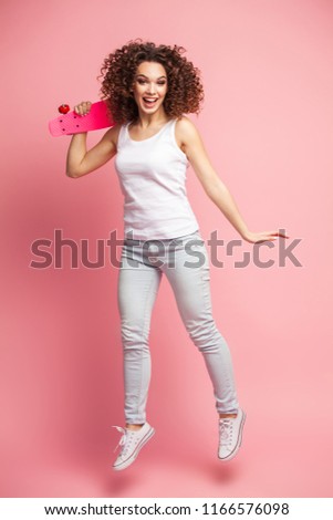 Full length portrait of a beautiful happy woman in summer clothes posing and looking away while jumping and holding skateboard isolated over pink background