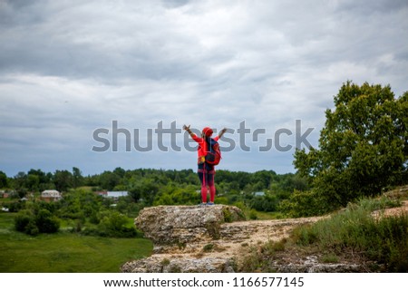 Photo from back of sportswoman with her hands up with backpack on top of hill against backdrop of picturesque countryside and gloomy sky