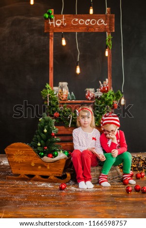 Blonde girl in res trousers and white blouse and blonde boy in red and white knitting hat sitting on the wooden floor with christmas balls
