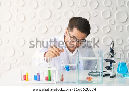 Scientist in White Gown and Protecting Glasses Drop Liquid from Test Tube to Fancy Fighting Fish in Glass Tank with Microscope, Conical Flask. Concept for Science, Education, Experiment, Research