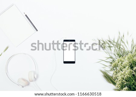 Blank smart phone near bouquet of white wildflowers, headphones and notepad, top view. Feminine working desk flat lay floral composition.