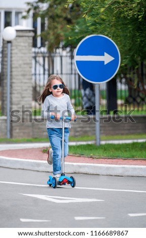 Happy beautiful girl is riding in the summer along the street on the kick scooter on a windy day. Summer break, baby having free time playing. The concept of a healthy lifestyle. Kids sport