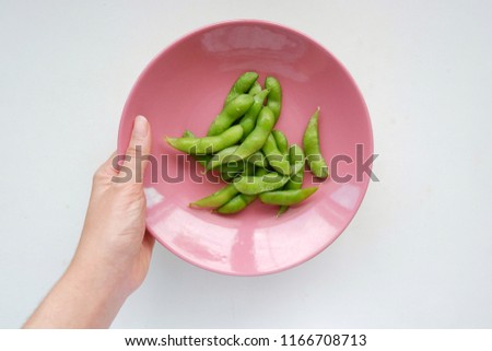 Top eye view closeups pieces of girl hand are holding many green Edamame soybean Japan in circle pastel pink color plate isolated on vintage white table background at a kitchen room.