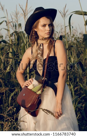 A young woman dressed as a cowgirl stands in a farmer's crop field corn at sunset and rests at a haystack