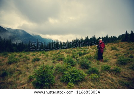 Adventurer with map is standing on the mountain meadow  on the background of low heavy clouds floating above green ridges stretching to the horizon - in mountains before storm. Back view. 
