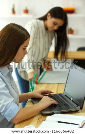 Portrait of two young female designers working on creating printout for clothes in bright modern office. One girl using laptop, sitting at desk, other measuring something on blurry background.