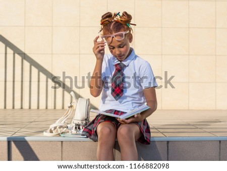 A girl in school uniform with a funny hairdo with colored pencils in her hair and with books sits on the steps in front of the school and reads the book, prepares for the lesson