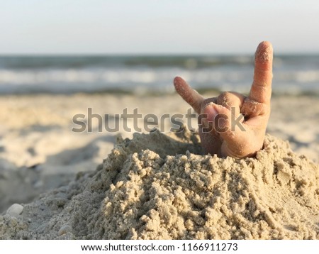 Rock and roll hand sign on a beach background. Human hand out of the hole on the sandy beach. Sea concept. 
