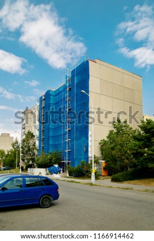 Thermal insulation, reconstruction work on a block of flats, blue canvas over scaffolding, street with cars, lamp, pavement, green tree, sunny blue sky, white clouds, vertical image