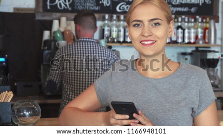 The woman is standing near the bar. She is checking her mobile phone while waiting for her order.The barista is making coffee for her. A woman comes to the cafe