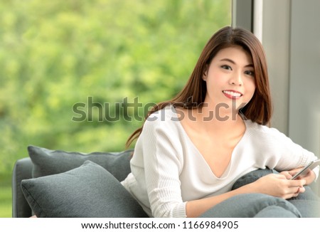 Close up shot portrait of young beautiful Asian woman sitting on sofa  and  relaxing manner and hold smarthphone in hand. Green nature blur in background with copy space on the left.