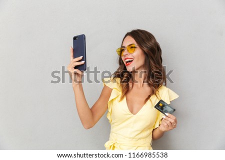 Portrait of an excited young girl in dress standing over gray background, taking selfie with mobile phone and showing plastic credit card